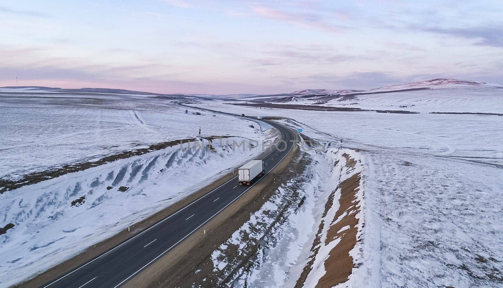 A lone semi truck drives along a winding rural highway through a snow-covered landscape during dusk in winter.
