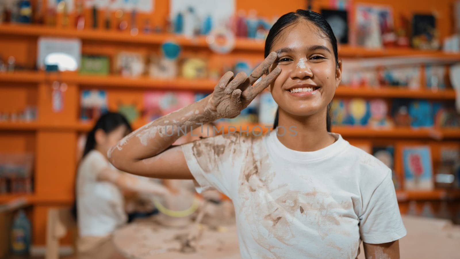 Happy caucasian girl pose at camera while diverse children modeling clay behind. Cute student wearing dirty shirt while looking at camera at workshop in art lesson. Blurring background. Edification.