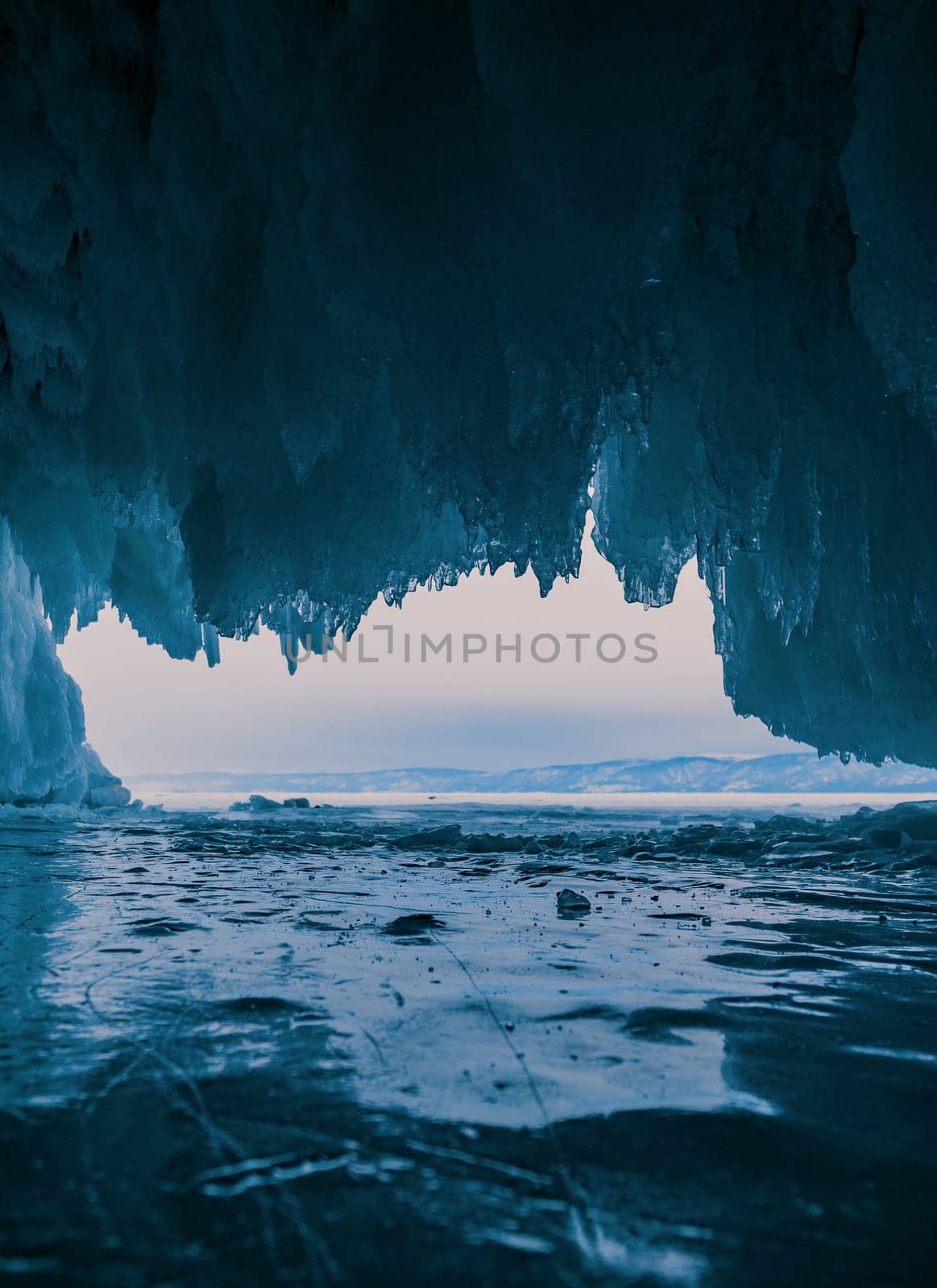 Inside a stunning ice cave on Lake Baikal, large icicles hang from the ceiling, creating a breathtaking winter landscape. Snow-covered mountains can be seen far in the distance.