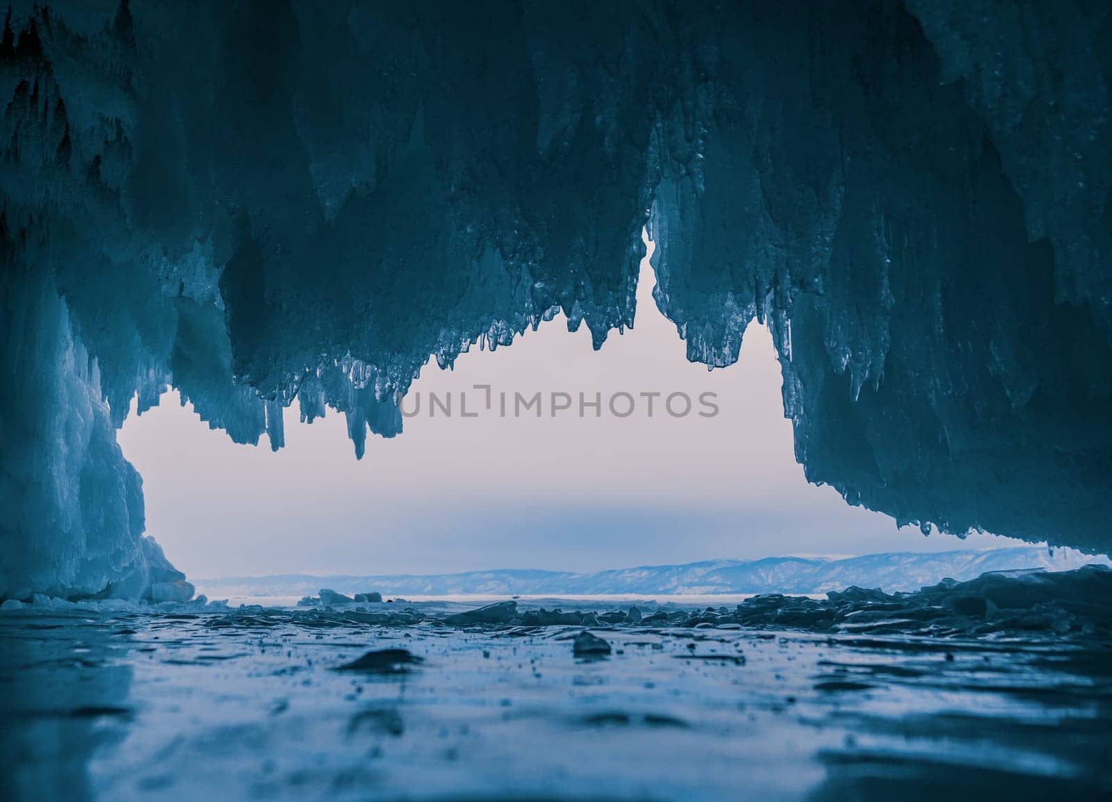 Inside a stunning ice cave on Lake Baikal, large icicles hang from the ceiling, creating a breathtaking winter landscape. Snow-covered mountains can be seen far in the distance. by Busker