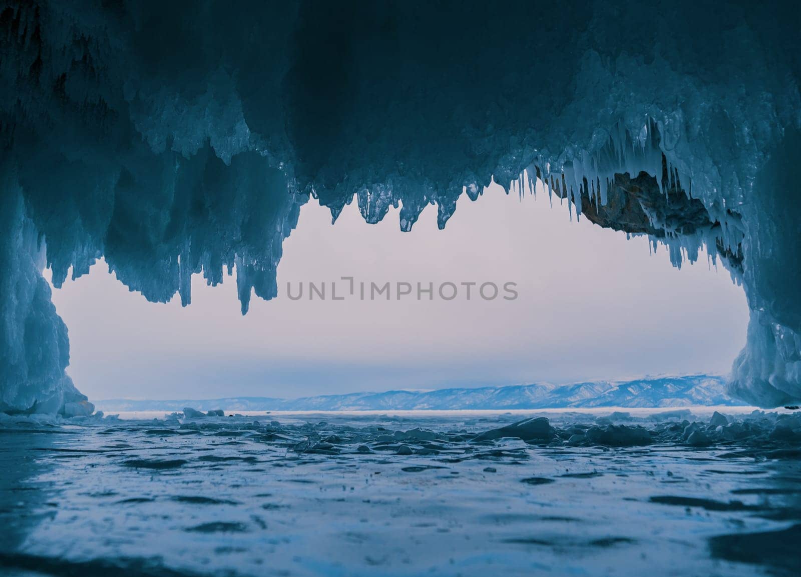 Inside a stunning ice cave on Lake Baikal, large icicles hang from the ceiling, creating a breathtaking winter landscape. Snow-covered mountains can be seen far in the distance.