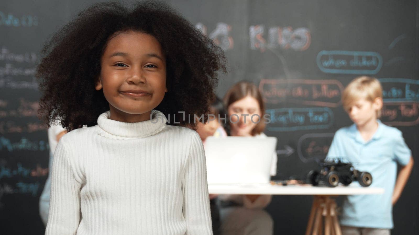 African girl smiling and looking at camera while multicultural friends working or learning engineering code or prompt in STEM technology classroom from young beautiful caucasian teacher. Erudition.