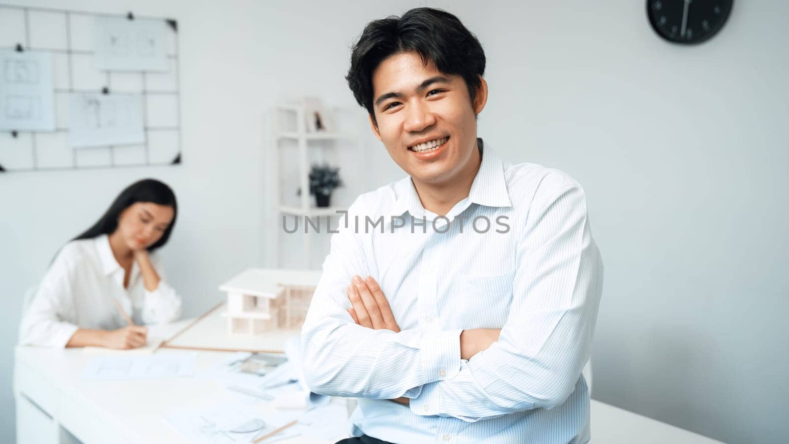 Professional asian businessman crosses arms with confident while young beautiful caucasian colleague drawing a blueprint on meeting table with house model and blueprint at modern office. Immaculate.