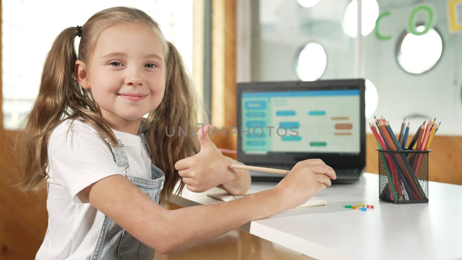 Little pretty caucasian girl smiling to camera while showing a thumb up gesture. Young child wearing headphone and casual dress while laptop display coding system or programing prompt. Erudition.