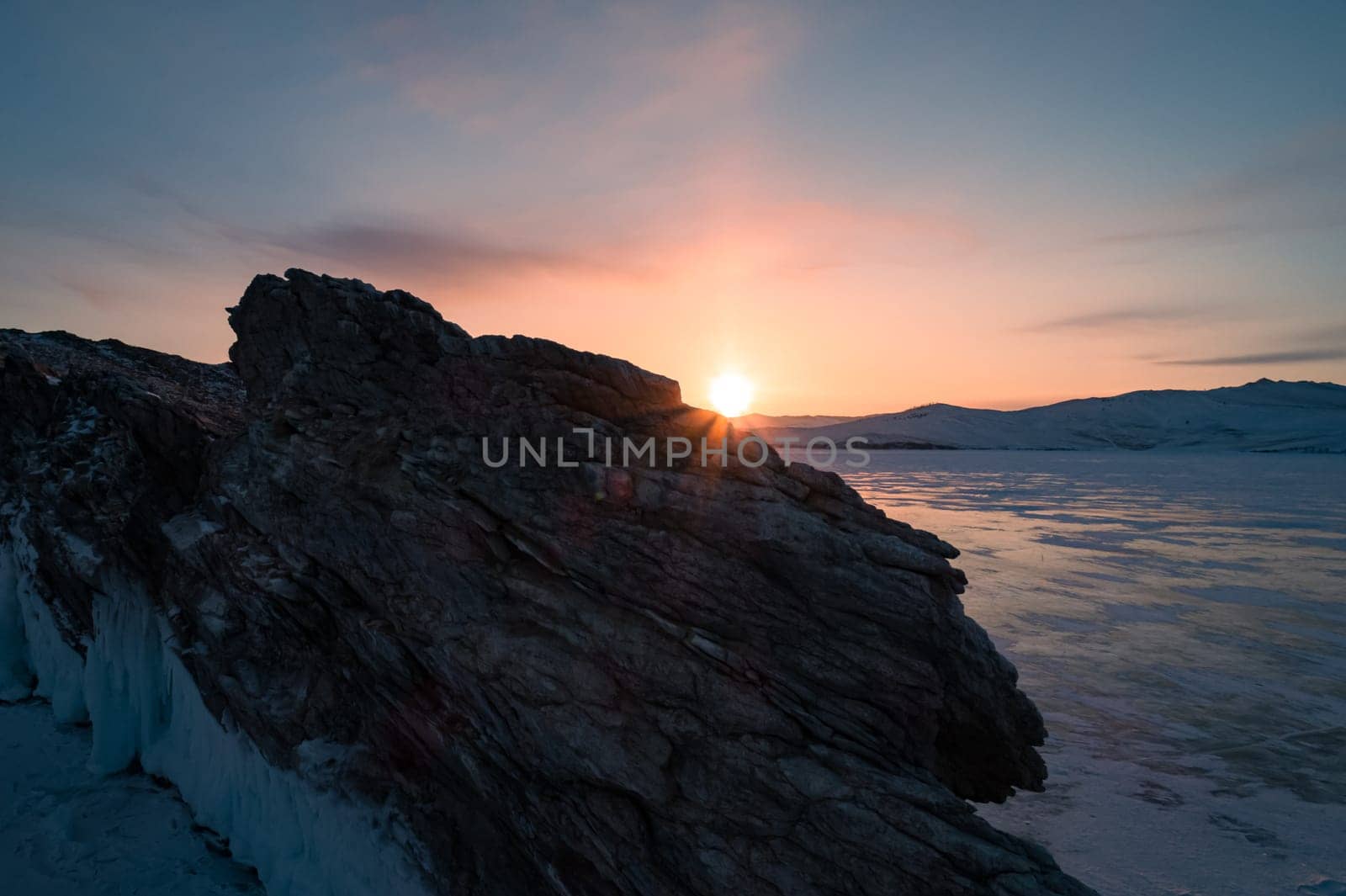 Aerial over the rocky island in lake Baikal. Winter landscape of frozen Baikal at beautiful orange sunrise. Sun reflections on the ice. Popular tourist spot by Busker