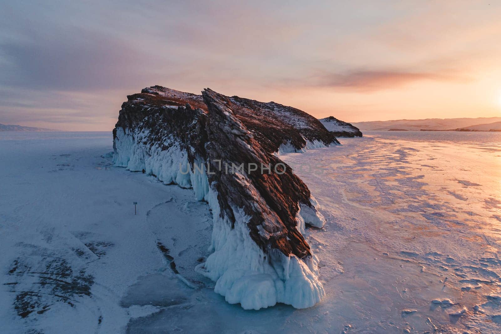 Aerial over the rocky island in lake Baikal. Winter landscape of frozen Baikal at beautiful orange sunrise. Sun reflections on the ice. Popular tourist spot.