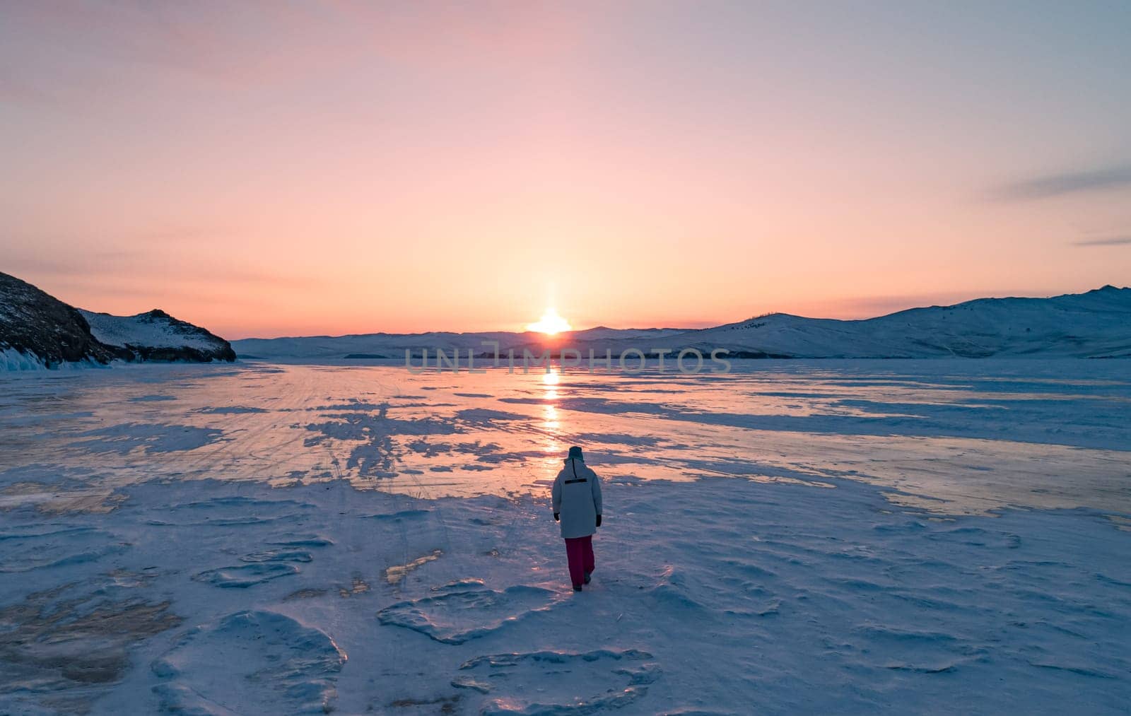 Aerial view on the young woman walking on the blue cracked ice of Baikal at beautiful orange sunrise. Sun reflections on the ice. Winter landscape of frozen Baikal by Busker