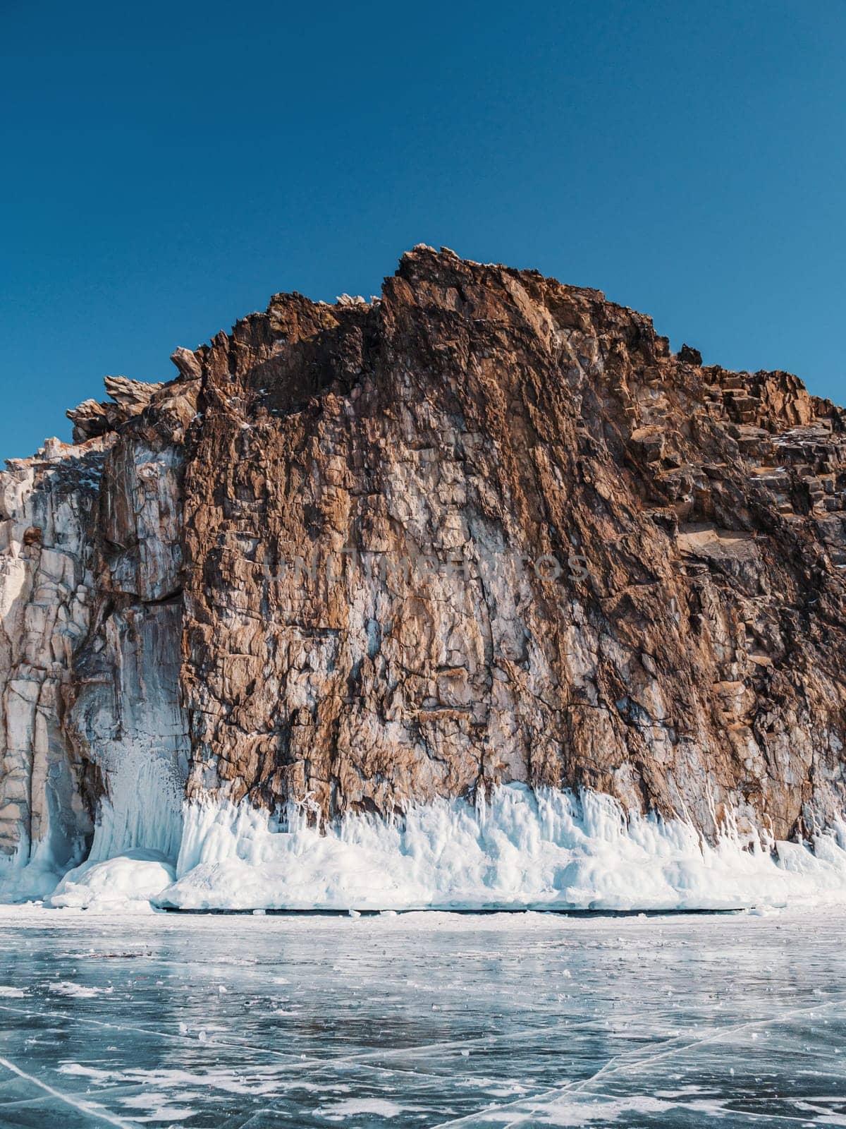 A majestic rocky cliff towers over the frozen surface of Lake Baikal, adorned with stunning ice formations and illuminated under a clear blue sky. by Busker