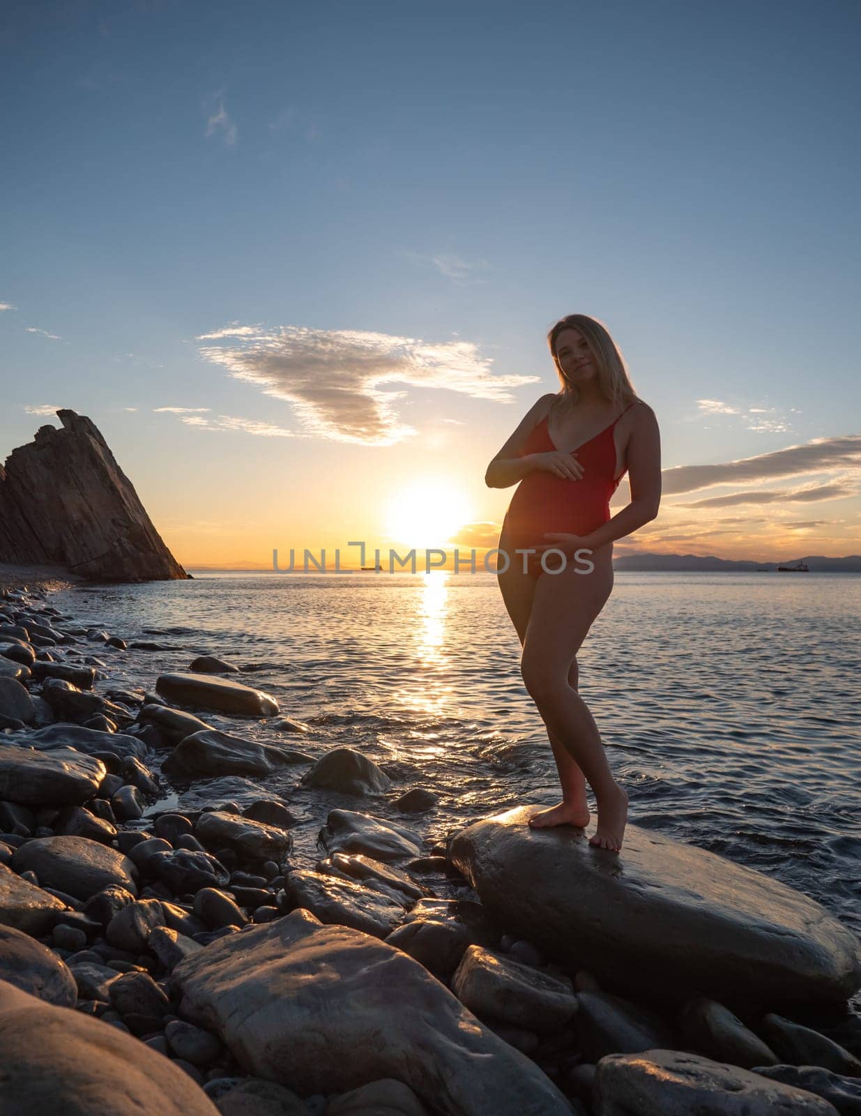 A pregnant woman stands on a rock by the seaside during sunrise, lovingly cradling her baby bump.