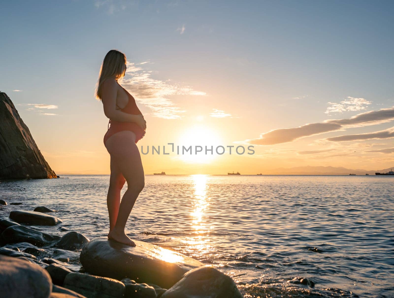 A pregnant woman stands on a rock by the seaside during sunrise, lovingly cradling her baby bump.