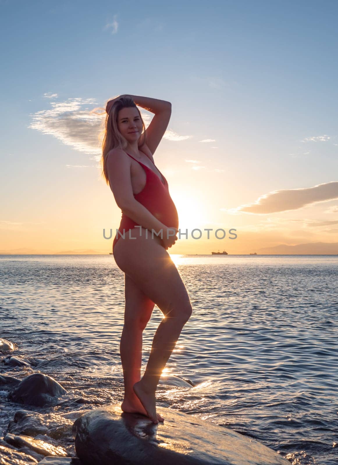 A pregnant woman stands on a rock by the seaside during sunrise, lovingly cradling her baby bump.