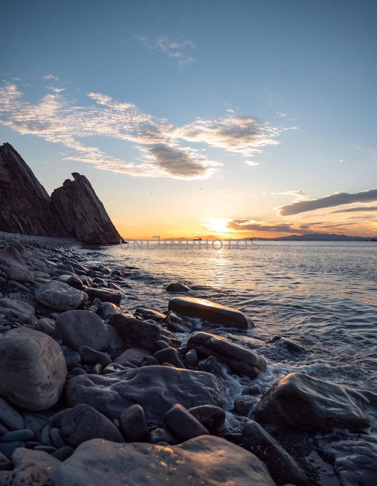 Sunset over rocky beach with silhouetted cliffs and calm ocean waters by Busker