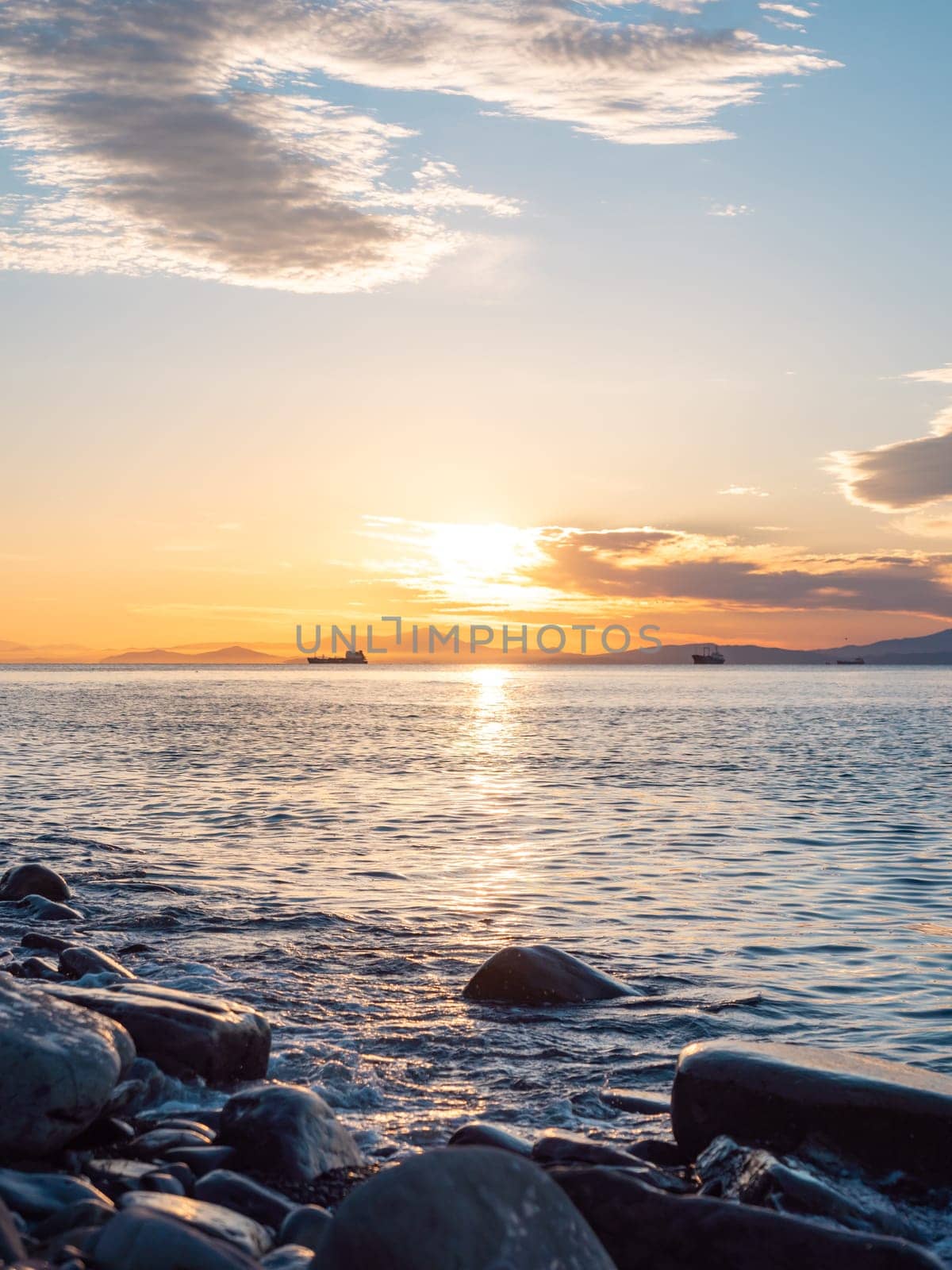 A tranquil sunset casts warm hues over a rocky beach and silhouetted cliffs. The calm ocean waters reflect the changing colors of the sky, creating a serene and picturesque coastal scene.