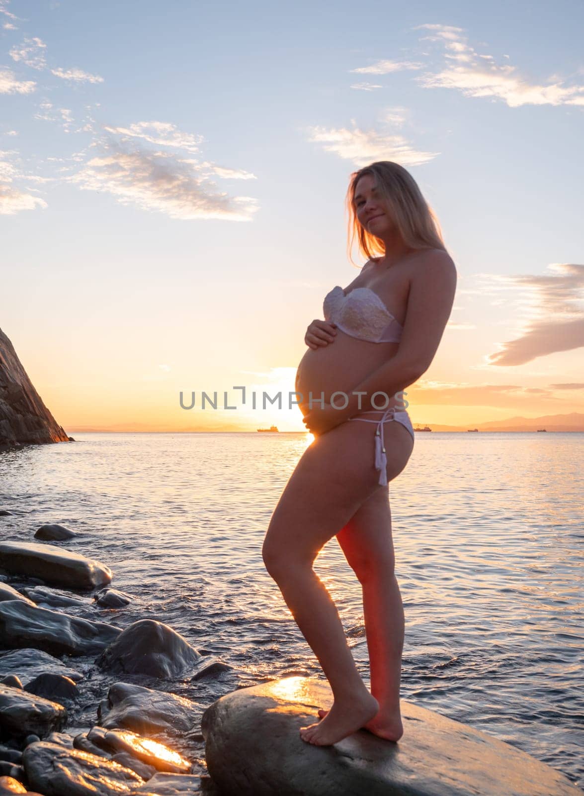 A pregnant woman in a white bikini stands on a rocky beach at sunrise, gently holding her belly. The background features calm water and a distant mountain range.