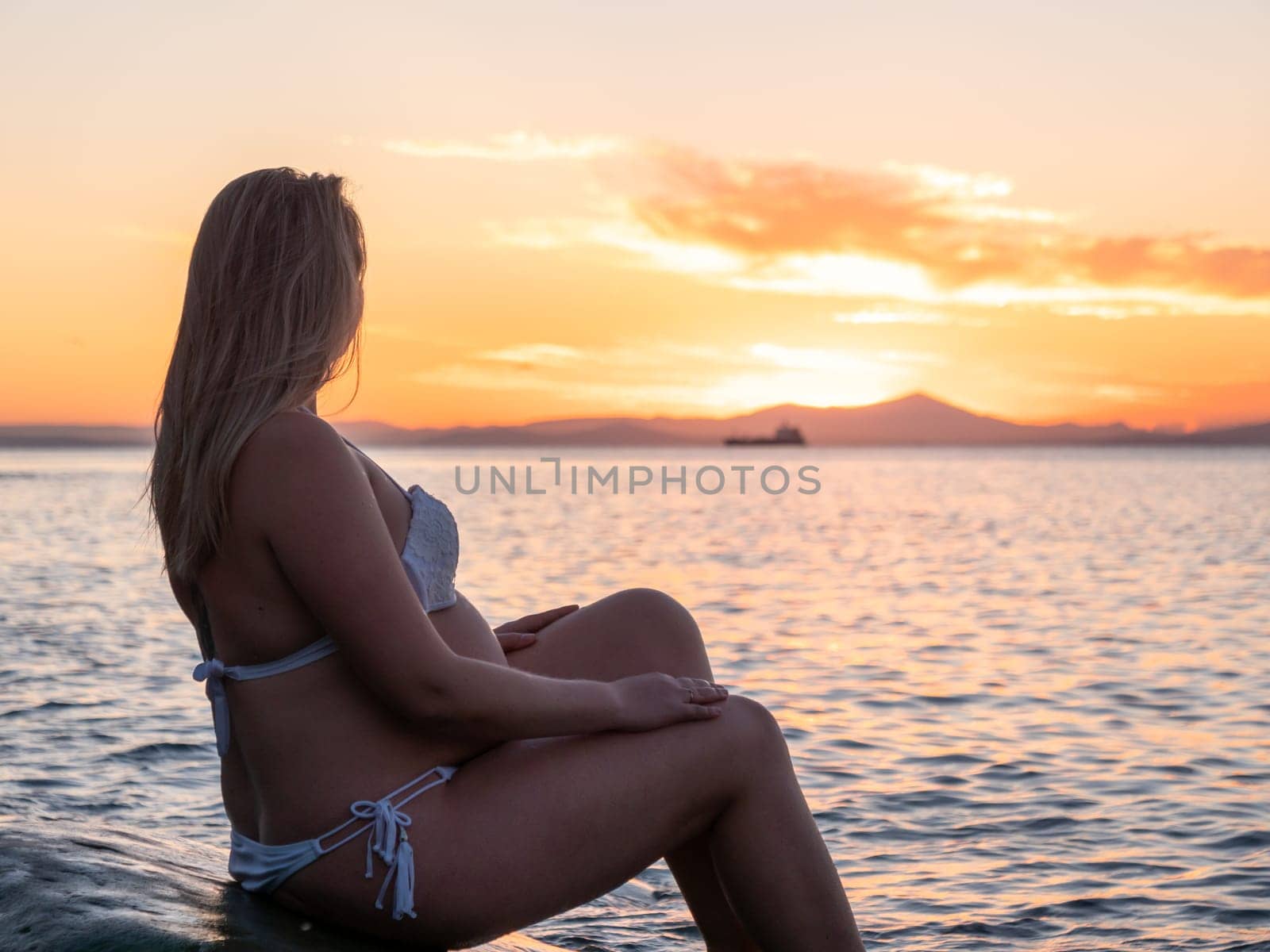 A pregnant woman in a white bikini sits on a rocky beach at sunrise. The background features calm water and a distant mountain range.