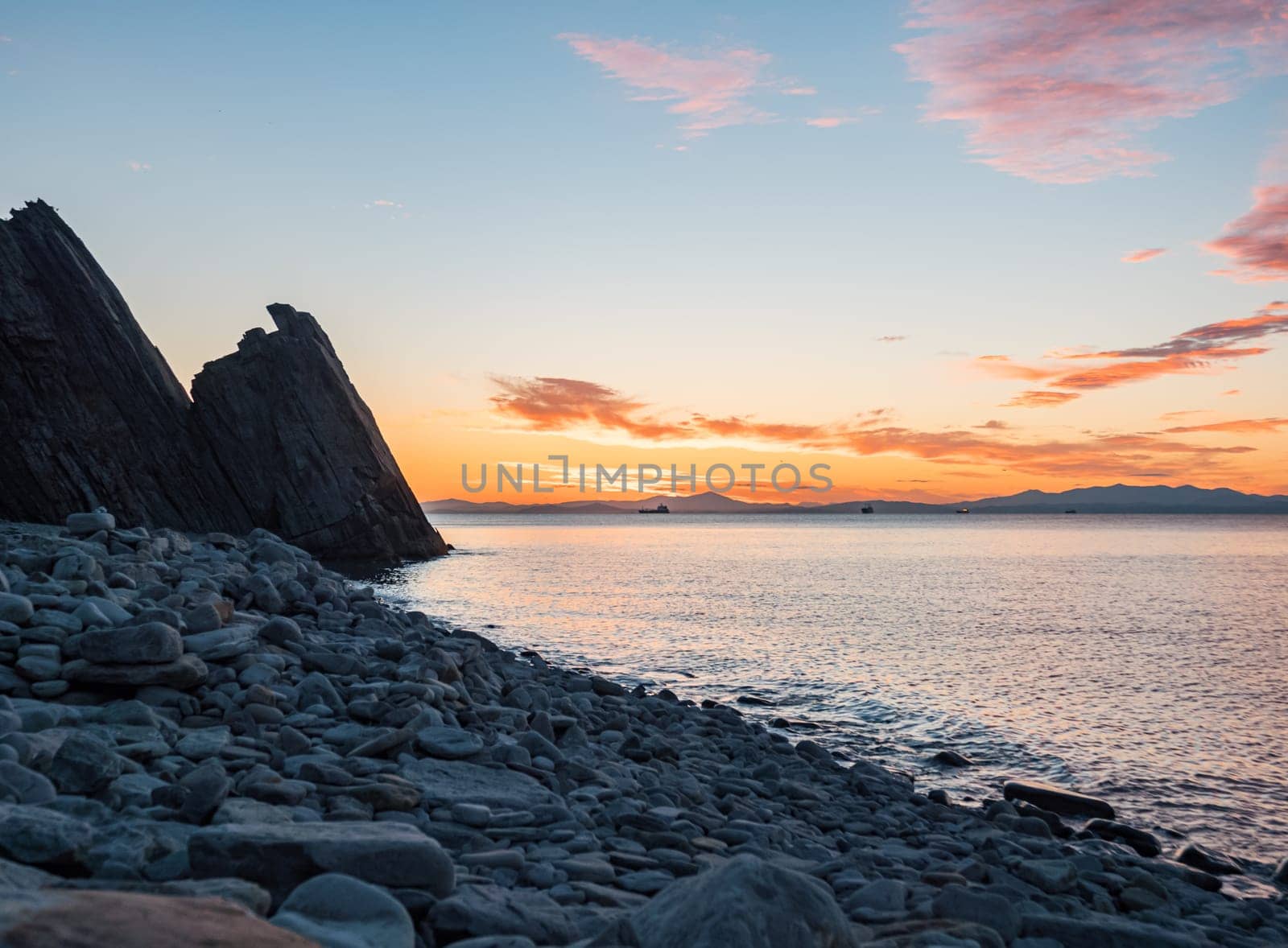 A tranquil sunset casts warm hues over a rocky beach and silhouetted cliffs. The calm ocean waters reflect the changing colors of the sky, creating a serene and picturesque coastal scene.