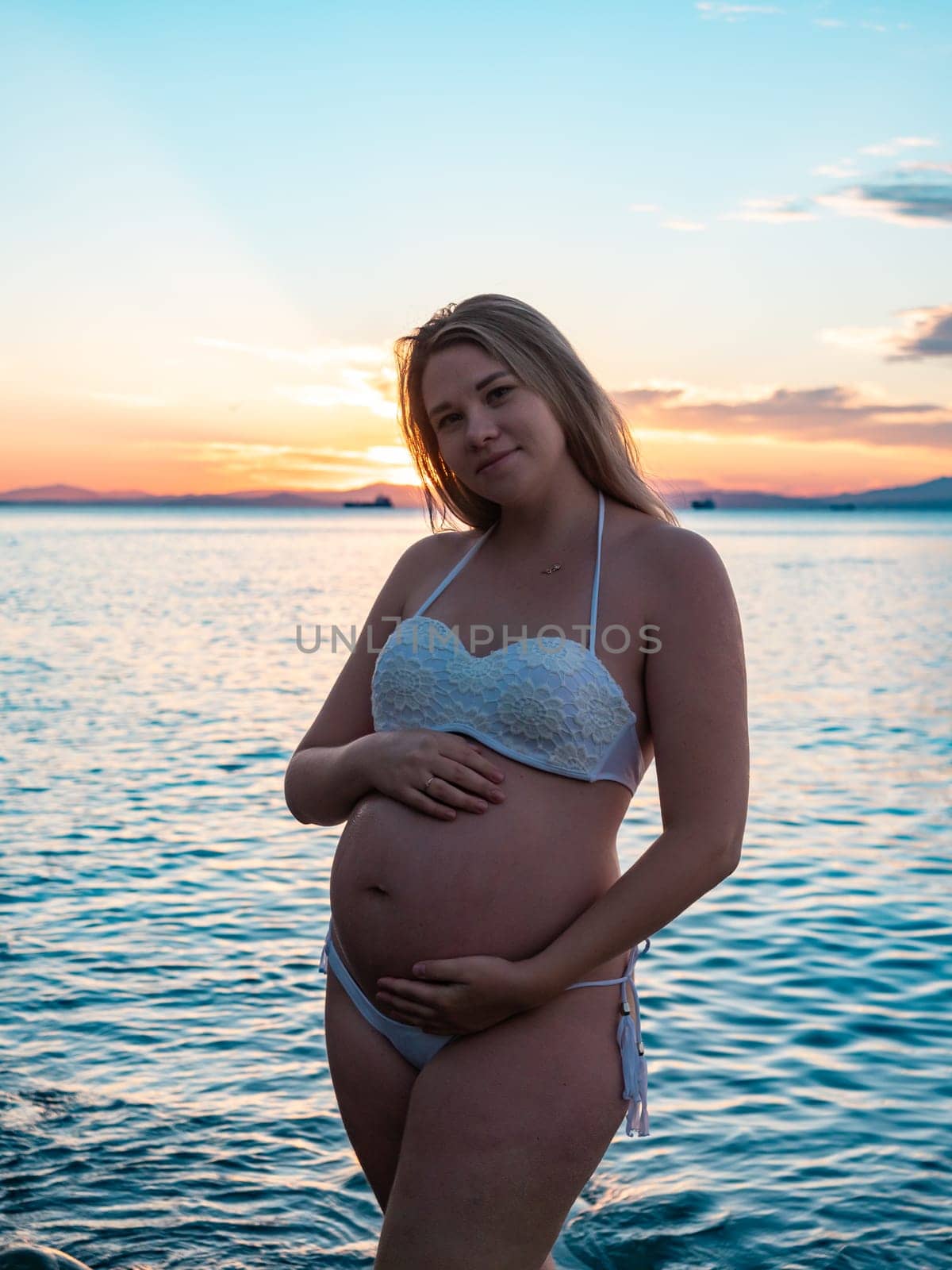 Pregnant woman in bikini posing on rocky beach at sunrise with mountain view by Busker