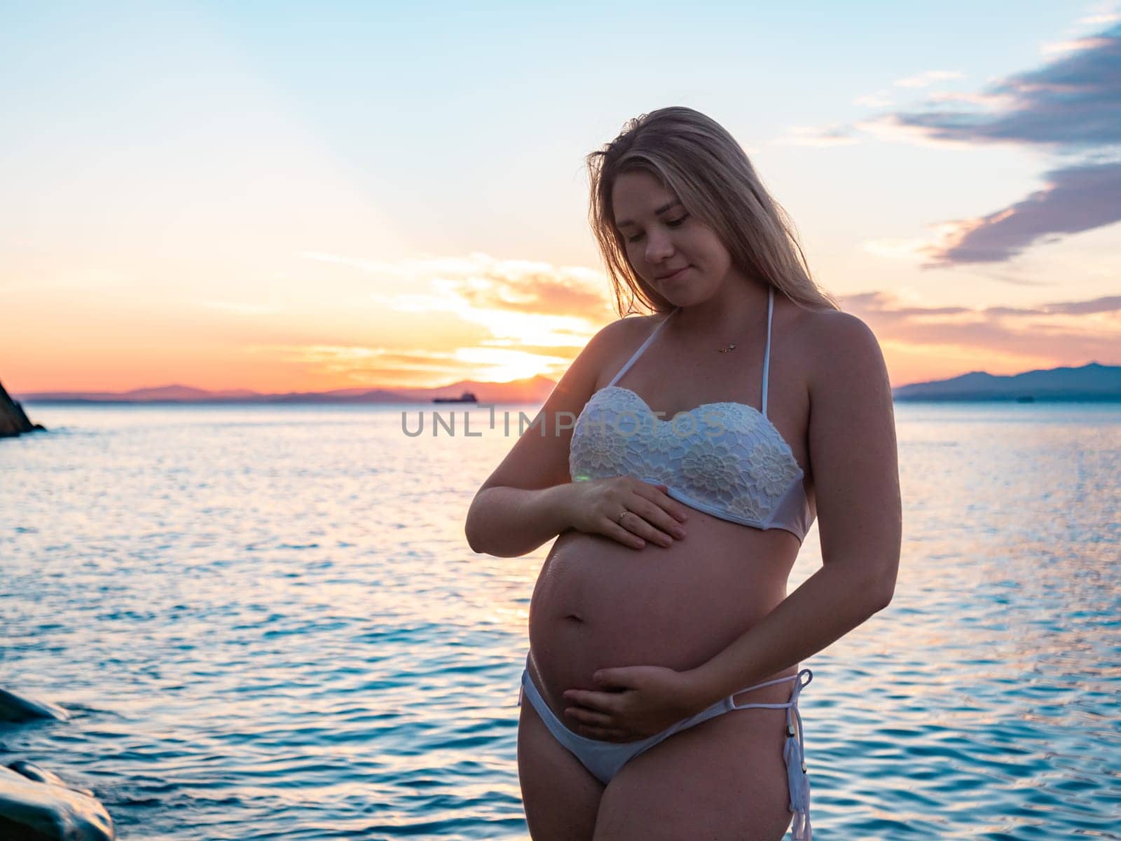 A pregnant woman in a white bikini stands on a rocky beach at sunrise, gently holding her belly. The background features calm water and a distant mountain range.