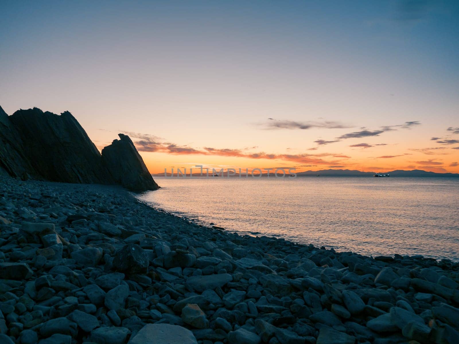 A tranquil sunset casts warm hues over a rocky beach and silhouetted cliffs. The calm ocean waters reflect the changing colors of the sky, creating a serene and picturesque coastal scene.