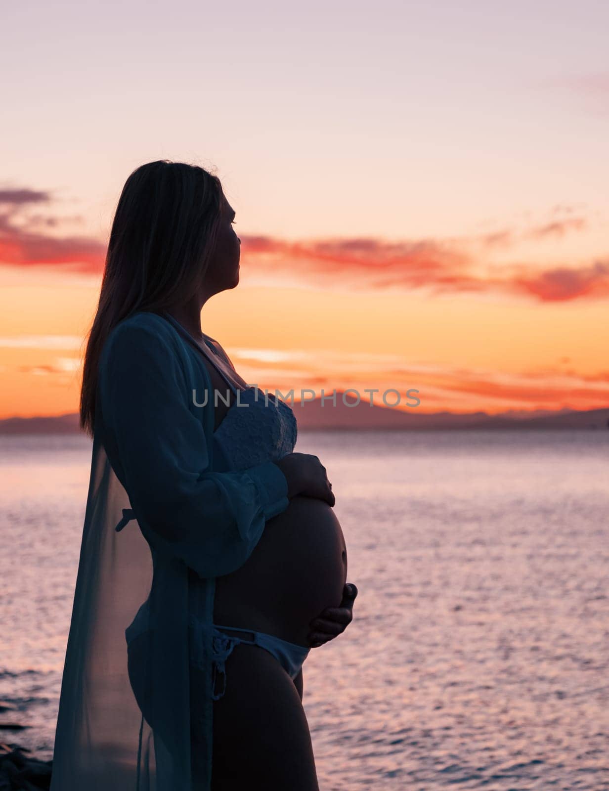 A pregnant woman in a white bikini stands on a rocky beach at sunrise, gently holding her belly. The background features calm water and a distant mountain range.