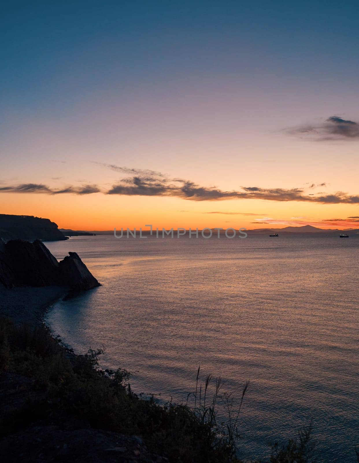 A tranquil sunset casts warm hues over a rocky beach and silhouetted cliffs. The calm ocean waters reflect the changing colors of the sky, creating a serene and picturesque coastal scene.