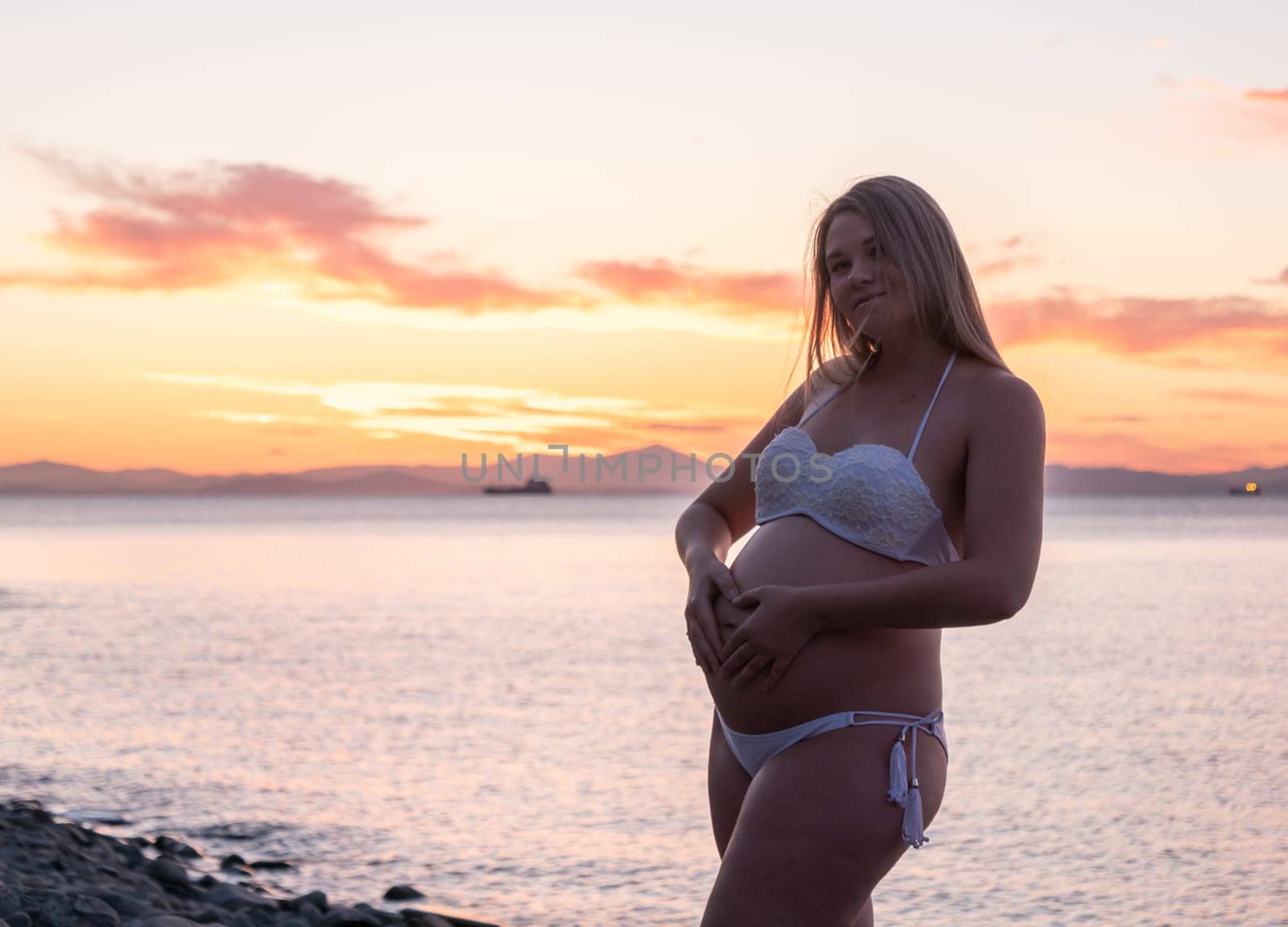 A pregnant woman in a white bikini stands on a rocky beach at sunrise, gently holding her belly. The background features calm water and a distant mountain range.