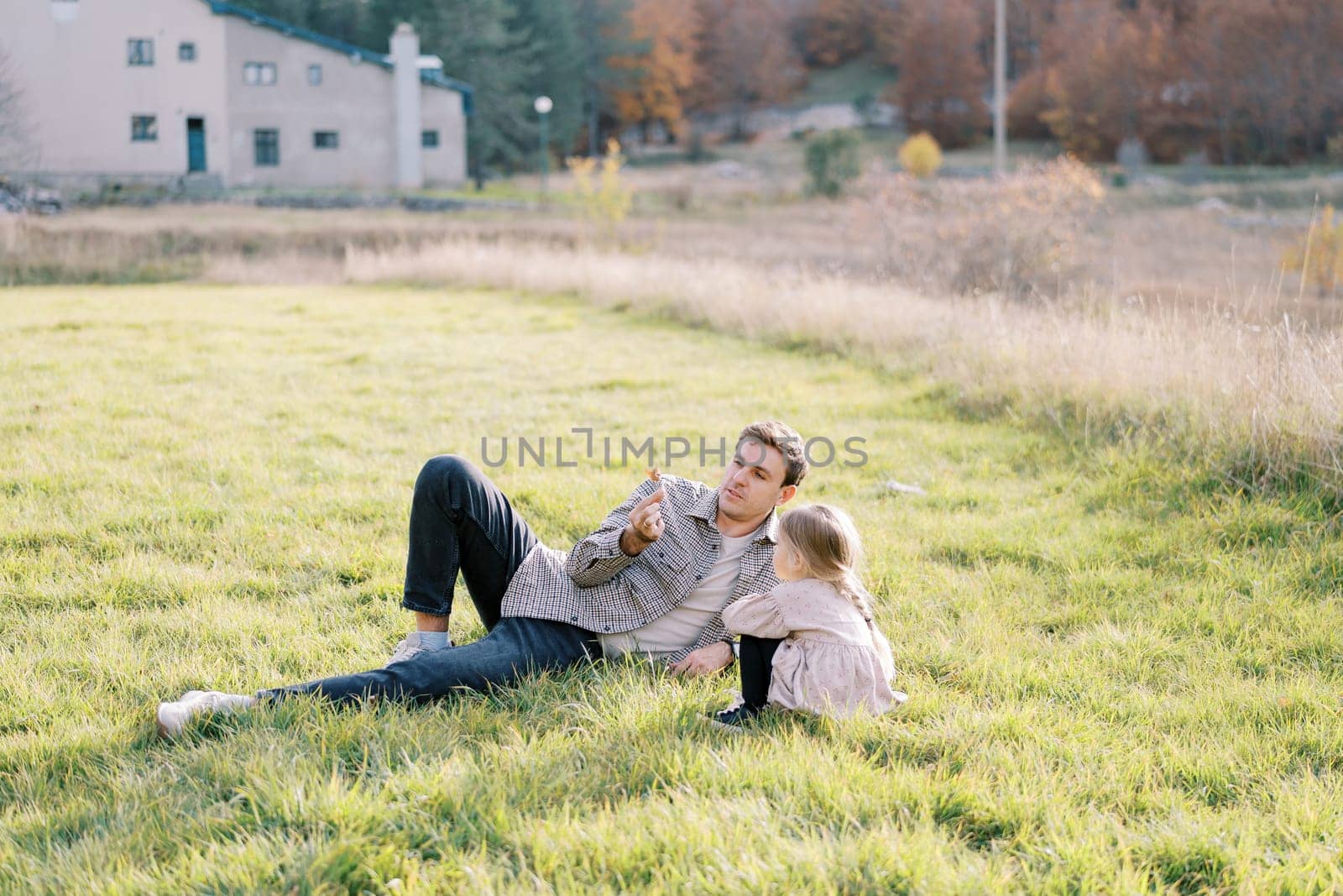 Little girl sits hugging her knees and looking at a dry leaf in the hands of her dad lying on the green lawn. High quality photo