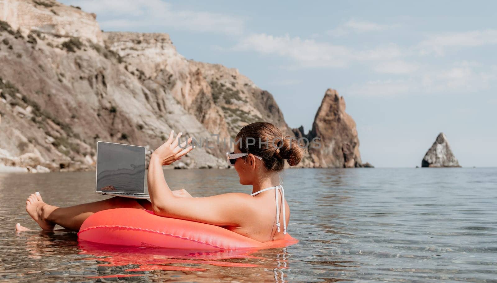 Woman freelancer works on laptop swimming in sea on pink inflatable ring. Pretty lady typing on computer while floating in the sea on inflatable donut at sunset. Freelance, remote work on vacation