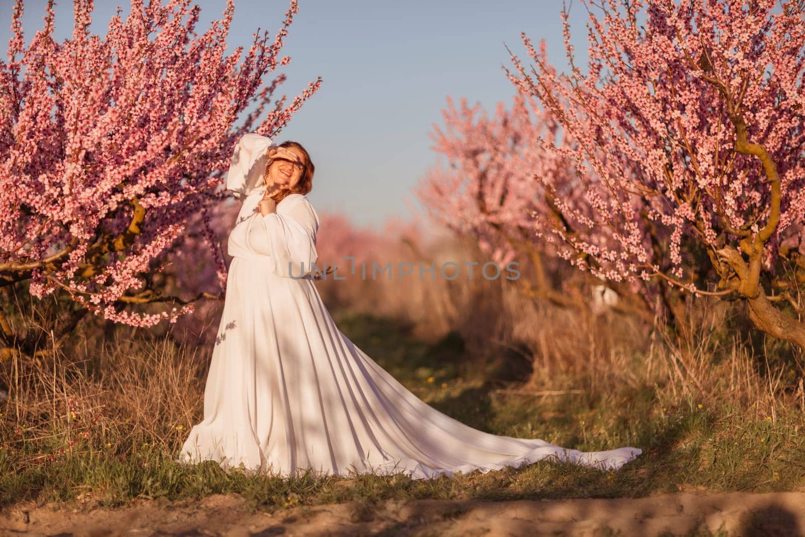 Woman blooming peach orchard. Against the backdrop of a picturesque peach orchard, a woman in a long white dress enjoys a peaceful walk in the park, surrounded by the beauty of nature. by Matiunina