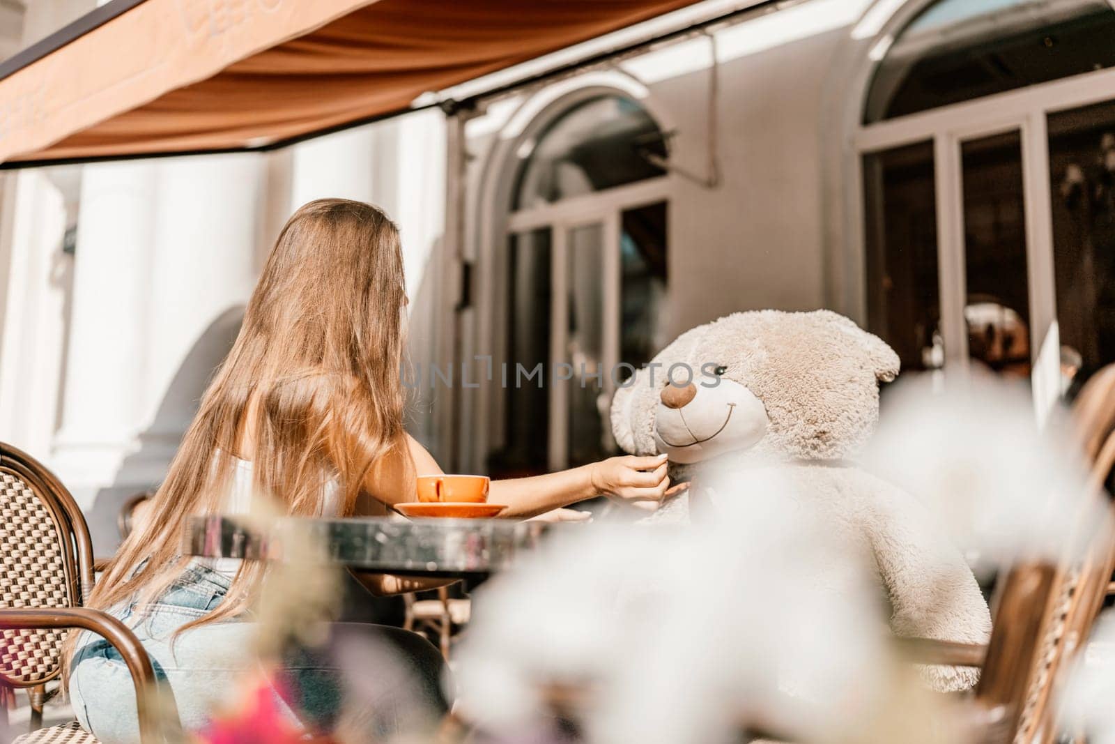 A woman sits cafe with a teddy bear next to her. The scene is set in a city with several chairs and tables around her. The woman is enjoying her time at the outdoor cafe