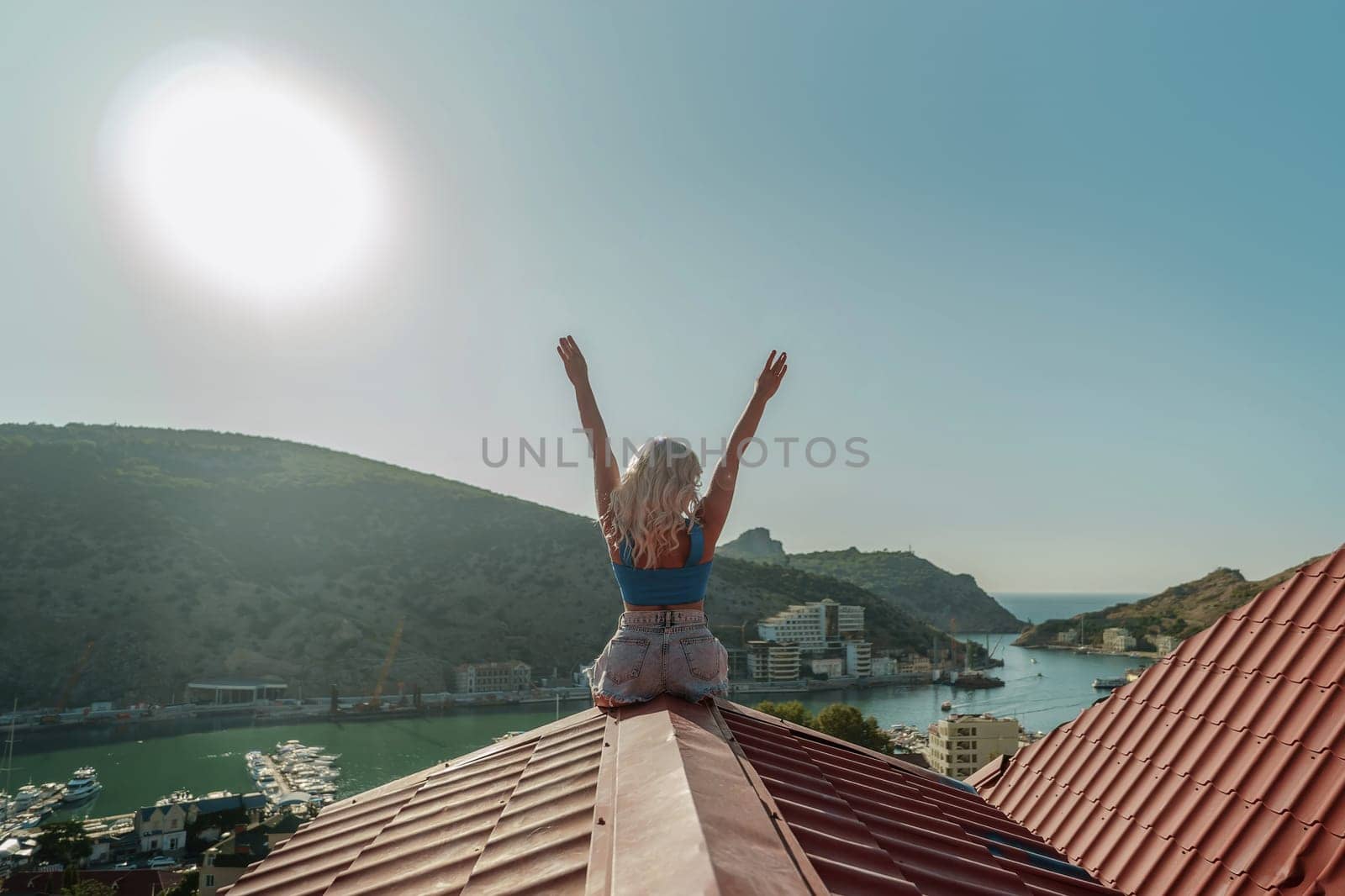 Woman sits on rooftop with outstretched arms, enjoys town view and sea mountains. Peaceful rooftop relaxation. Below her, there is a town with several boats visible in the water.