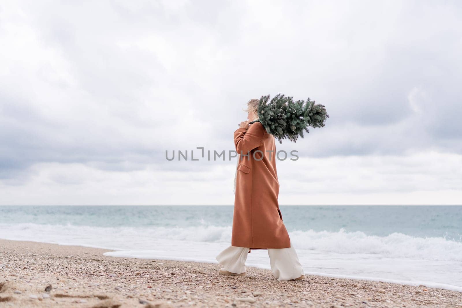 Blond woman Christmas sea. Christmas portrait of a happy woman walking along the beach and holding a Christmas tree on her shoulder. She is wearing a brown coat and a white suit