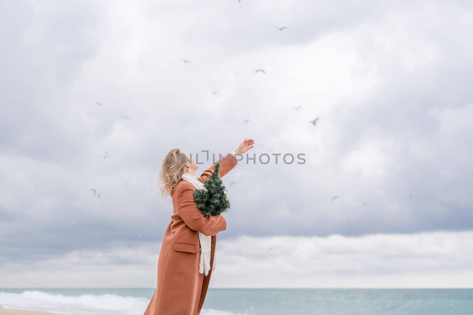 Blond woman Christmas sea. Christmas portrait of a happy woman walking along the beach and holding a Christmas tree on her shoulder. She is wearing a brown coat and a white suit