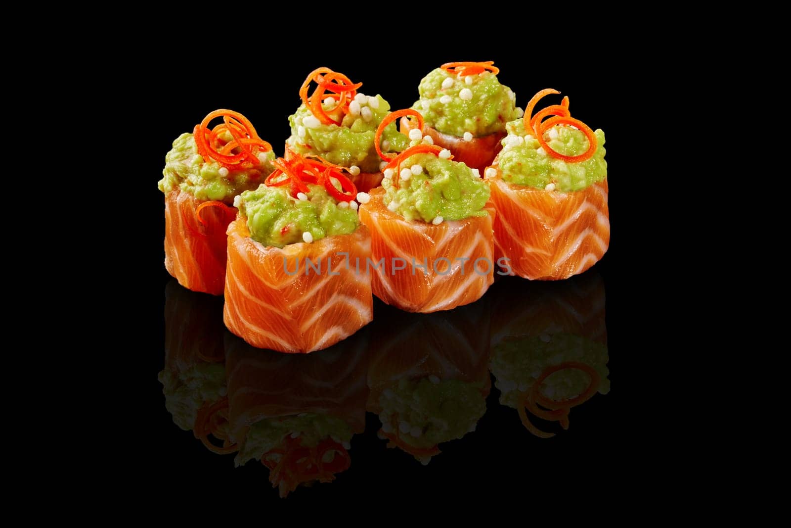 Colorful sushi rolls wrapped in raw salmon fillet slices topped with spicy avocado spread and crispy rice balls garnished with vegetable shavings on black glossy background. Popular Japanese dish