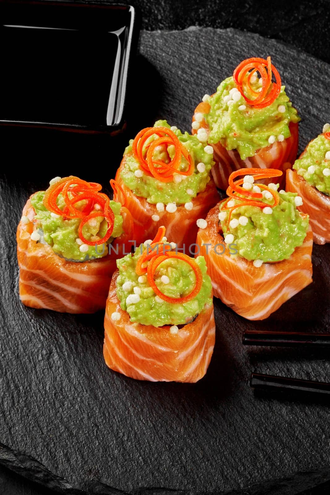 Colorful Japanese sushi rolls wrapped in raw salmon fillet slices topped with spicy avocado spread and crispy rice balls garnished with vegetable shavings served on black slate board with soy sauce