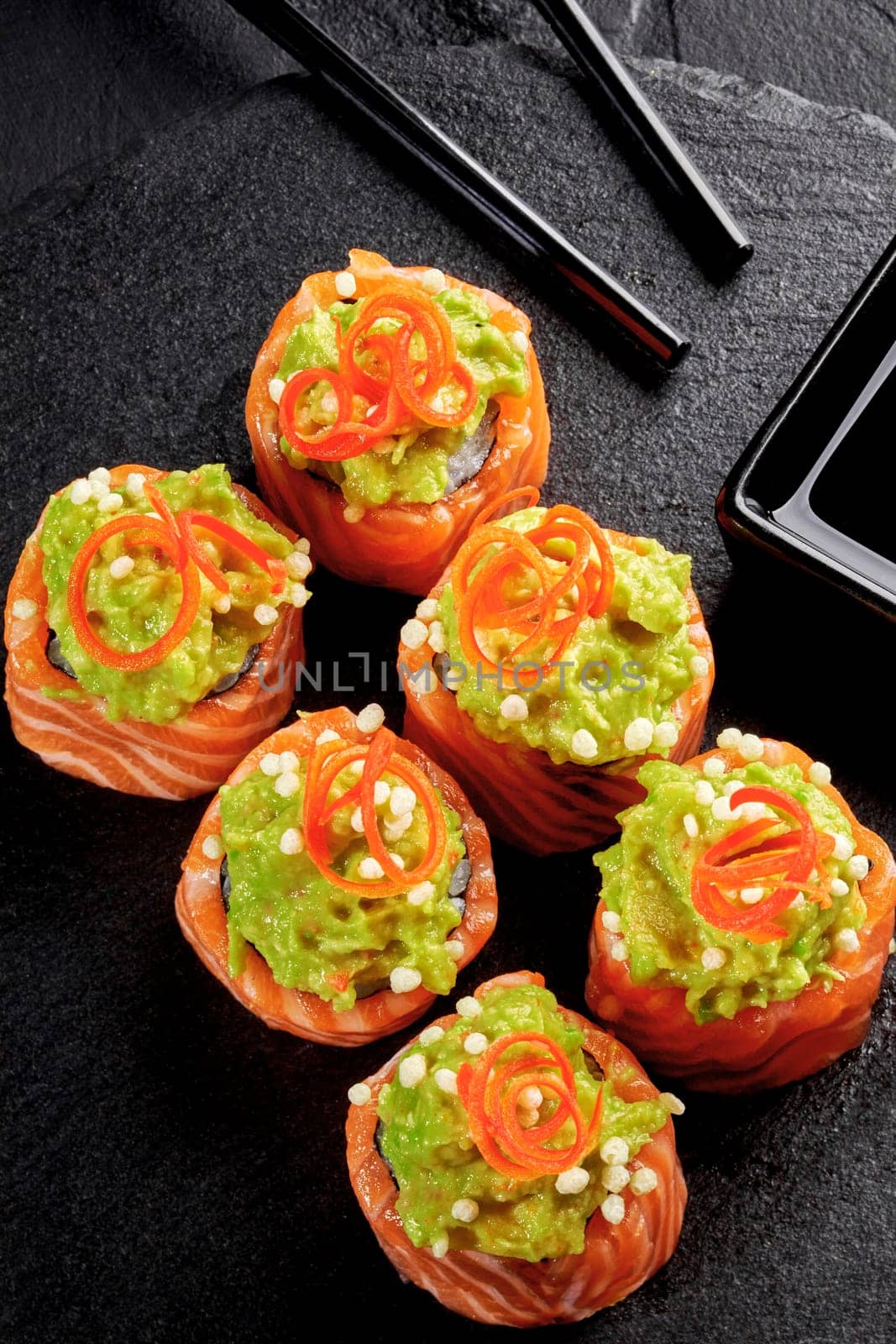 Artfully arranged salmon sushi topped with mashed avocado and delicate bell pepper curls, served on textured stone plate with soy sauce and chopsticks