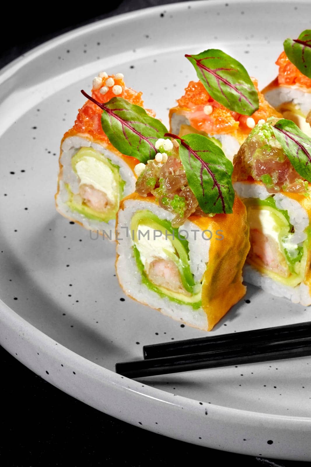 Artfully arranged colorful sushi rolls, wrapped in yellow mamenori, with shrimp tempura, cream cheese and avocado topped with salmon tartare placed on speckled ceramic plate with black chopsticks