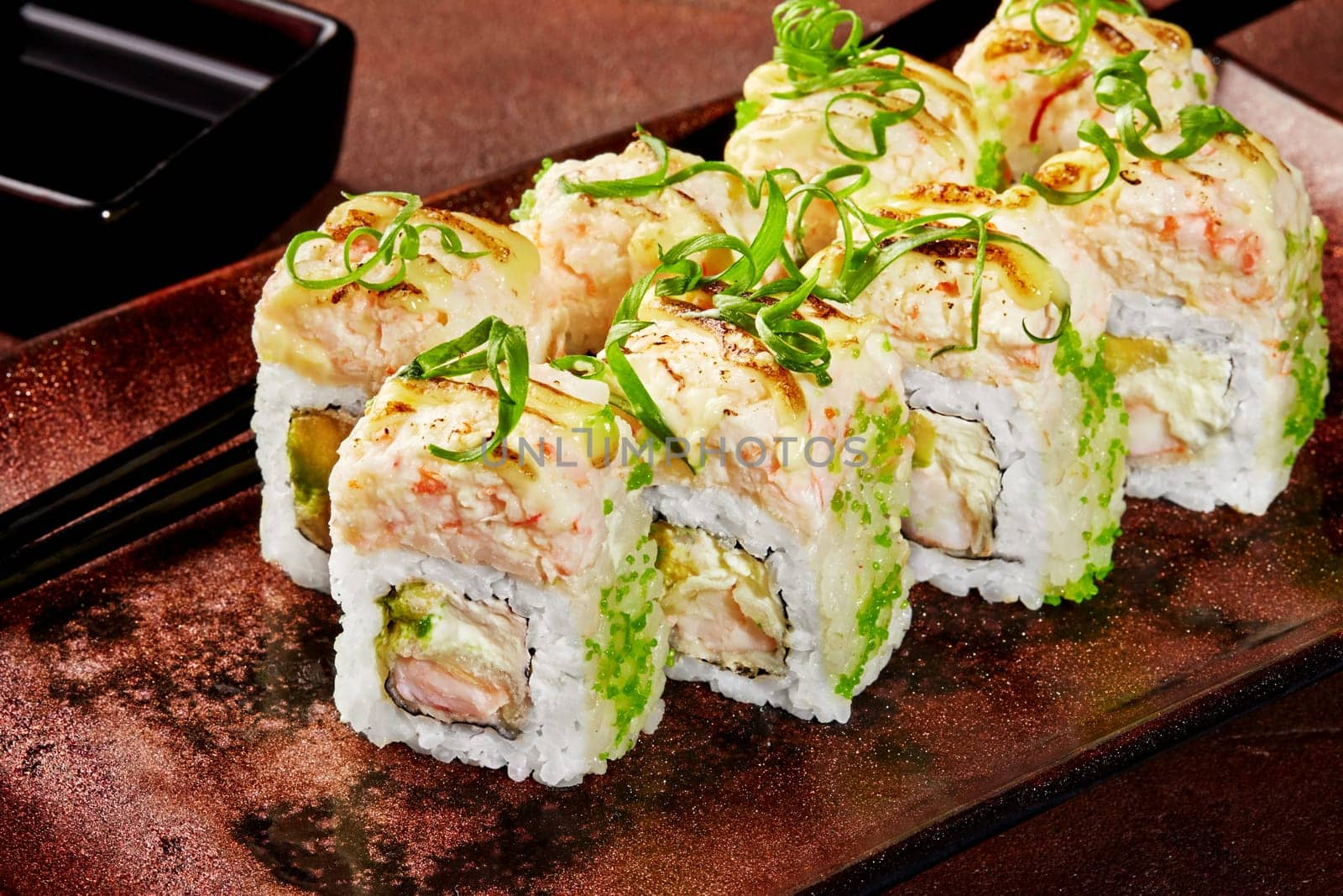 Close-up of uramaki sushi rolls coated with green tobiko filled with shrimp, avocado and cream cheese topped with spicy sauce and greens, traditionally served with soy sauce on ceramic plate