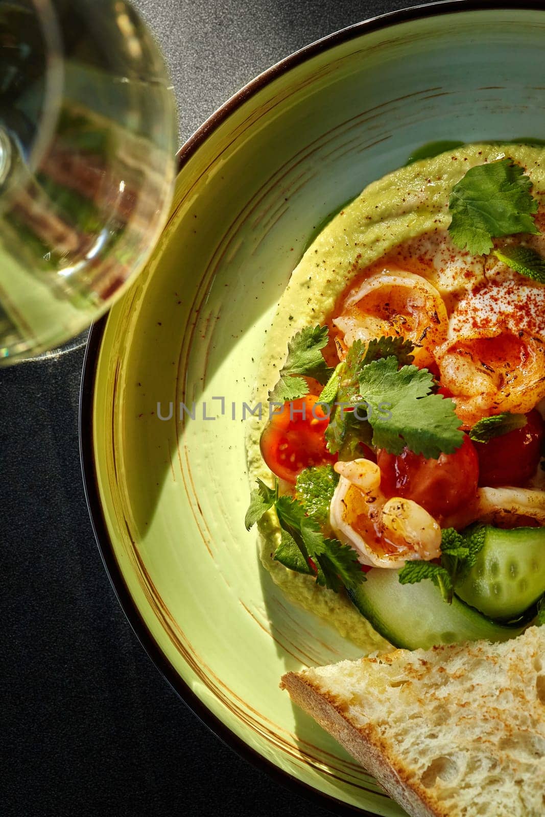 Delicate creamy hummus topped with succulent fried shrimps, fresh cherry tomatoes, cucumber slices and herbs served on ceramic plate with browned toast and glass of wine