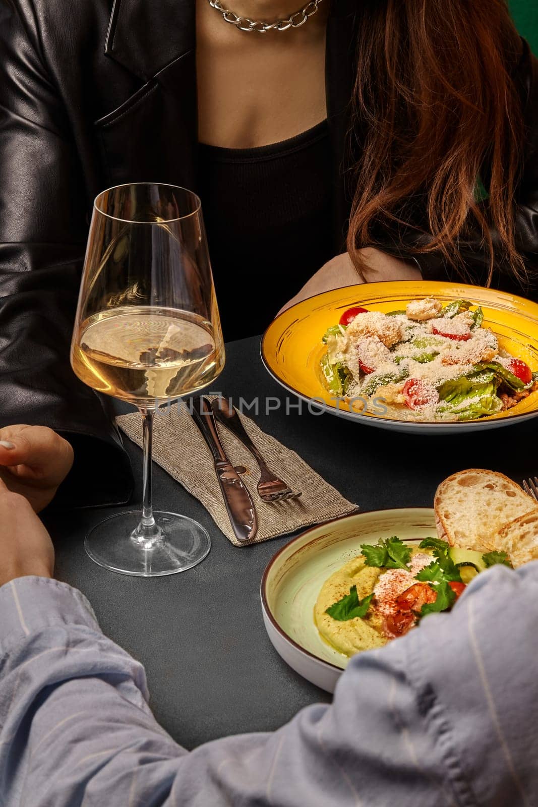 Elegant couple sharing moment over gourmet Caesar salad and creamy hummus with shrimps and greens, accompanied by glass of white wine, in stylish romantic dining setting