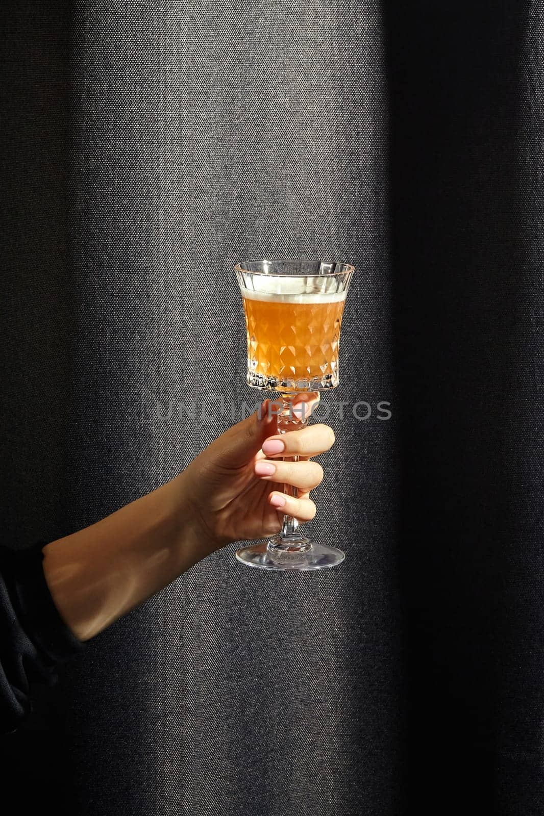 Female hand elegantly holding crystal glass filled with whiskey sour, cocktail in amber hue contrasting with dark textured fabric background