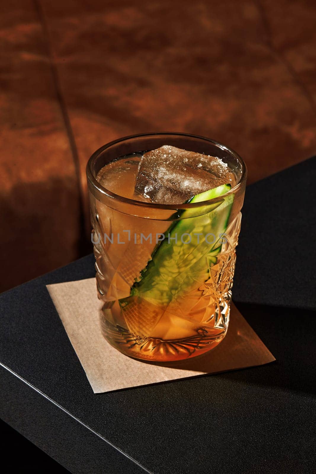 Glass of intriguing gin garden cocktail with translucent golden tone with ice cubes and fresh cucumber slice, presented on dark table, basking in warm light