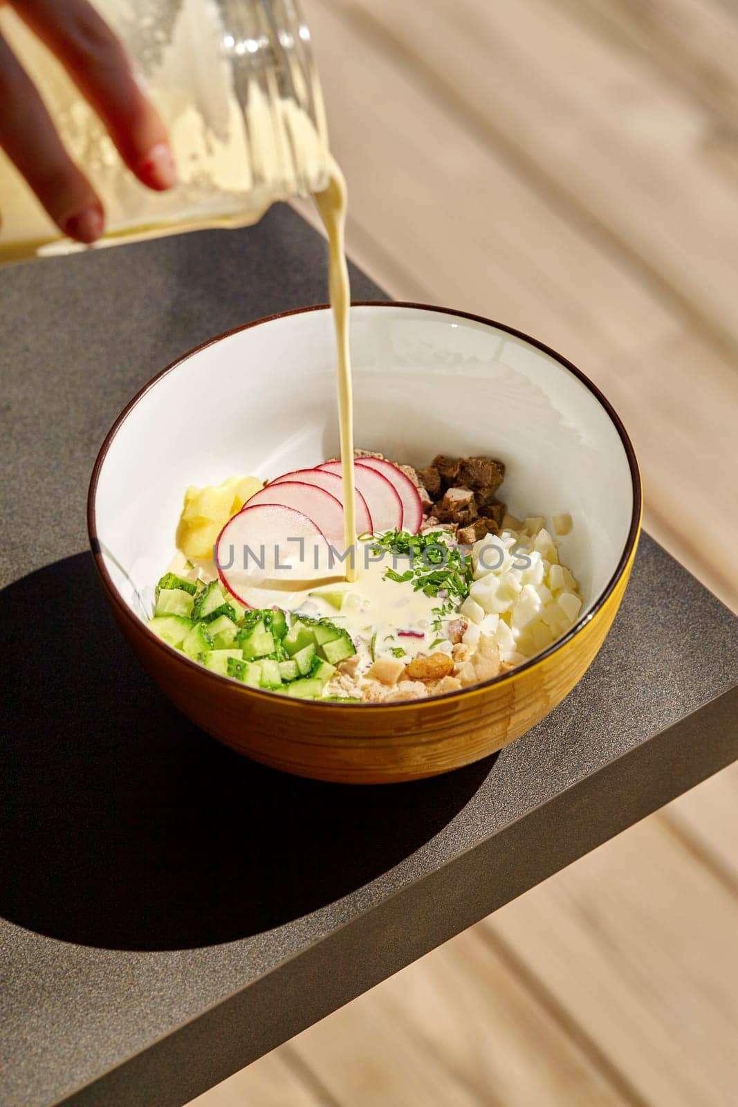 Final step in preparing classic Russian summer okroshka soup, with kefir poured over poured over chopped vegetables, eggs, and meats in ceramic bowl illuminated by sunlight