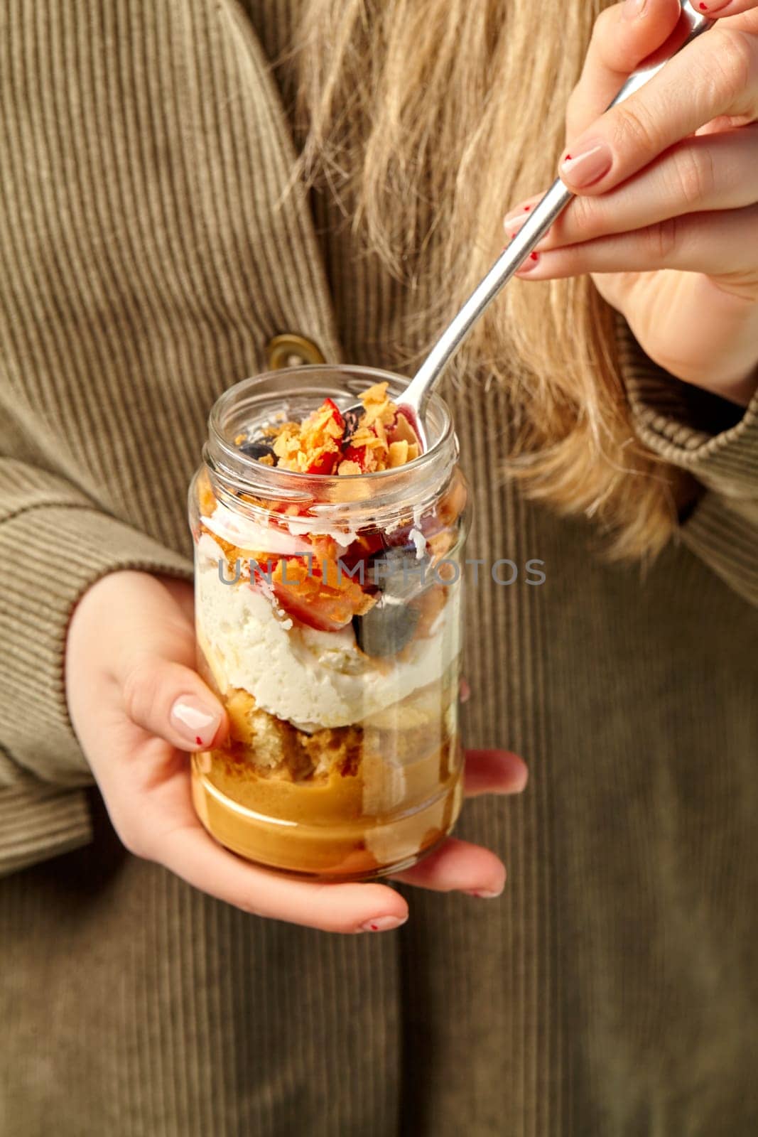 Woman scooping out of jar tender dessert of airy sponge cake soaked with classic Tres Leches sauce topped with rich Philadelphia cream cheese layer, fresh berries, and sprinkle of caramel crumble