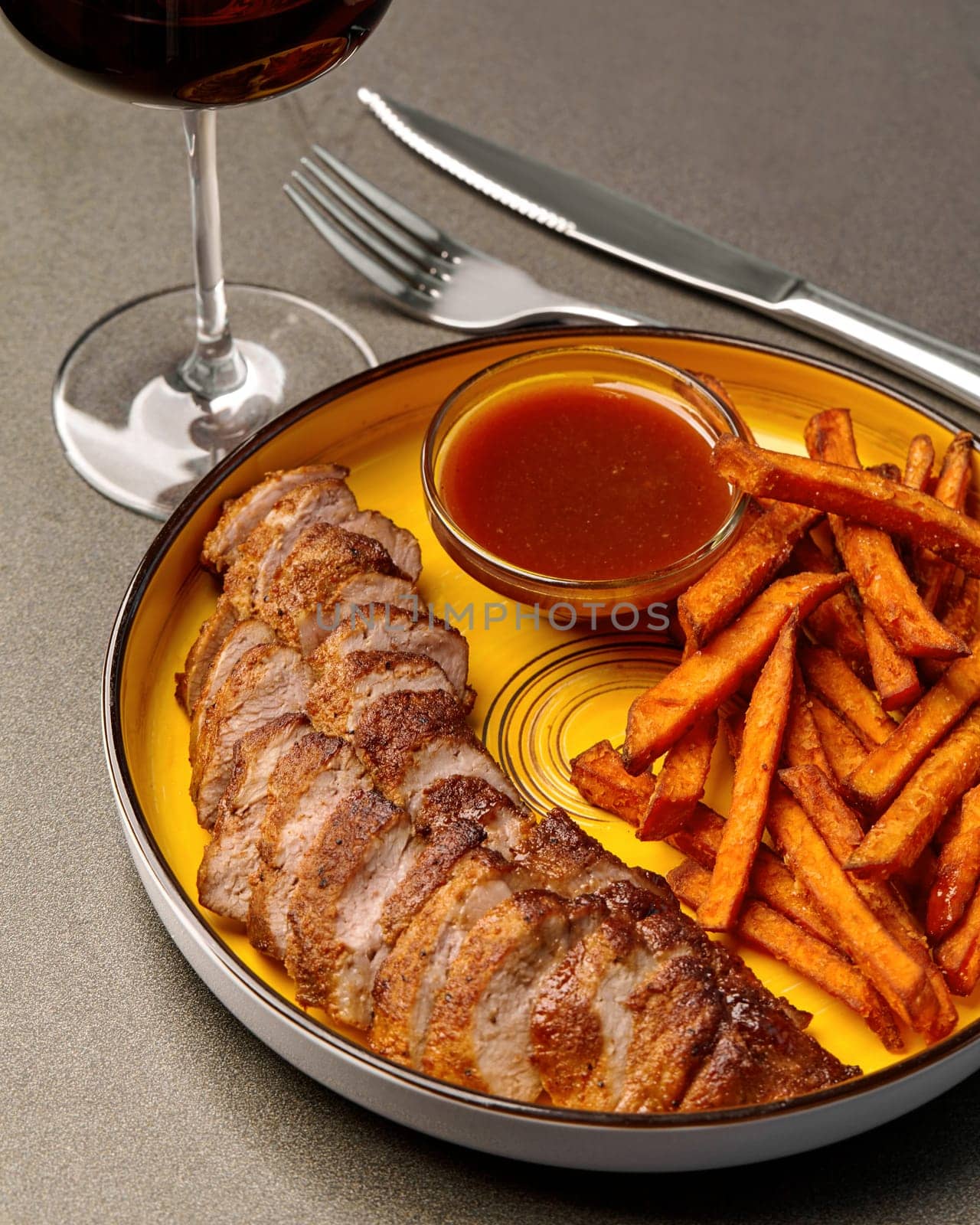 Grilled pork neck steak served on yellow plate with crispy sweet potato fries and side of tangy pineapple BBQ sauce, accompanied by glass of red wine