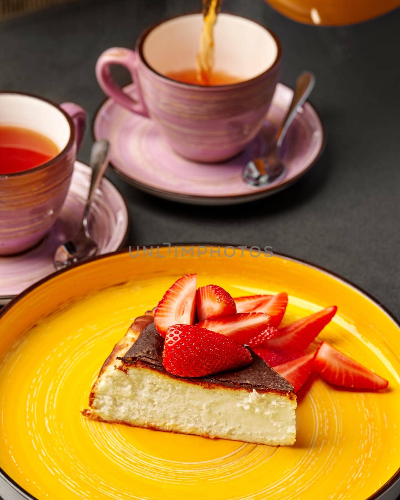 Inviting dessert setting featuring soft delicate cheesecake slice with fresh ripe strawberry on yellow plate with tea poured into cups, perfect for relaxing afternoon break