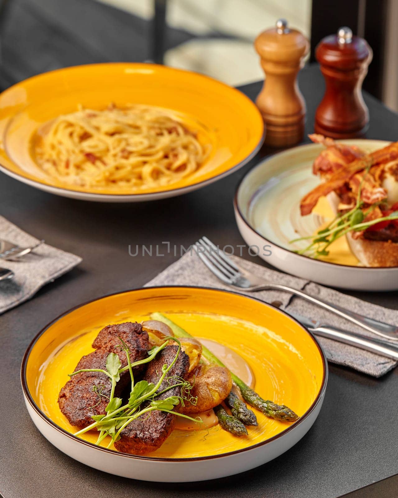 Warm inviting meal presented on vibrant yellow plates including pork steak with baked potatoes and asparagus, creamy pasta and bacon-topped bruschetta in background