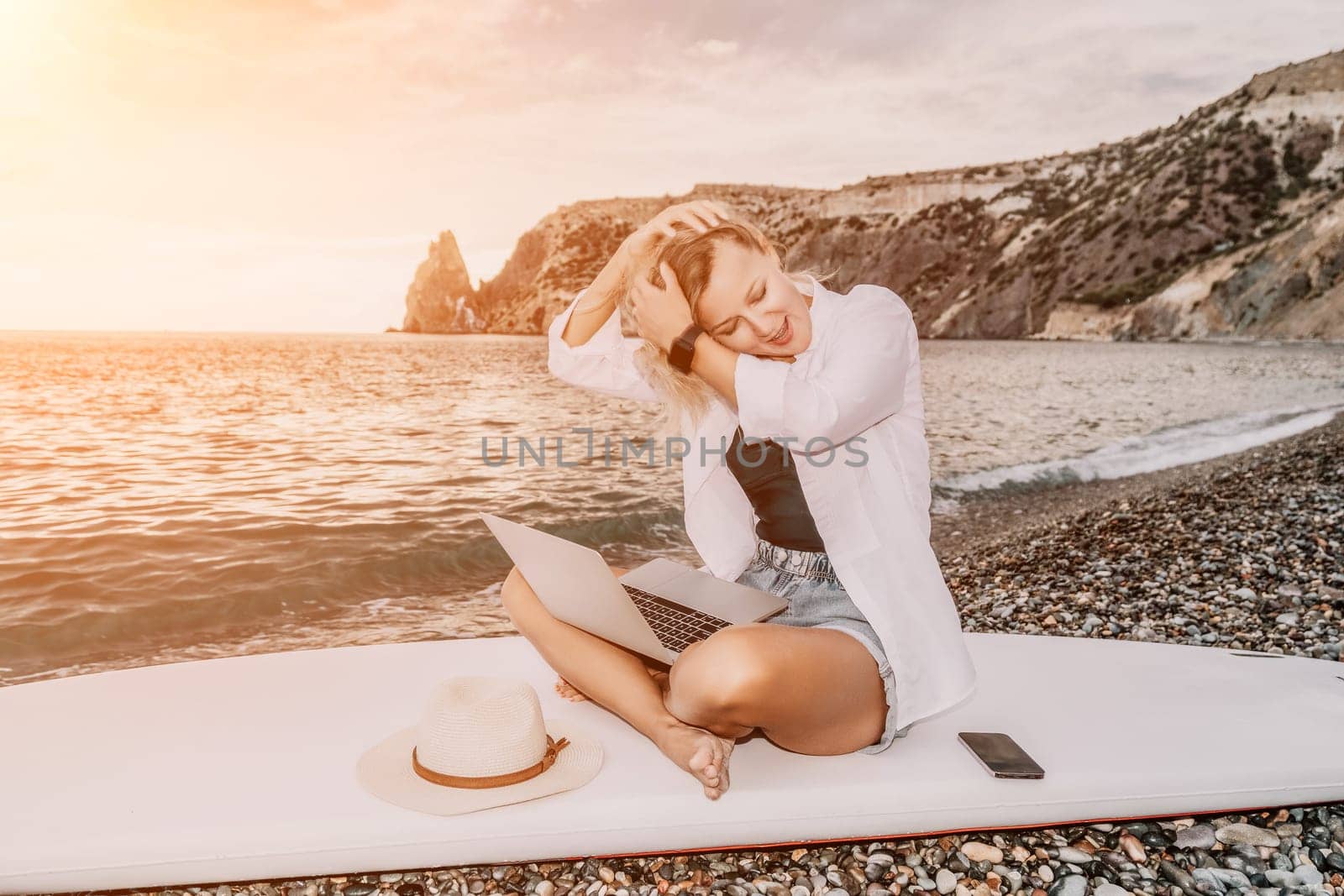 Successful business woman in yellow hat working on laptop by the sea. Pretty lady typing on computer at summer day outdoors. Freelance, travel and holidays concept.