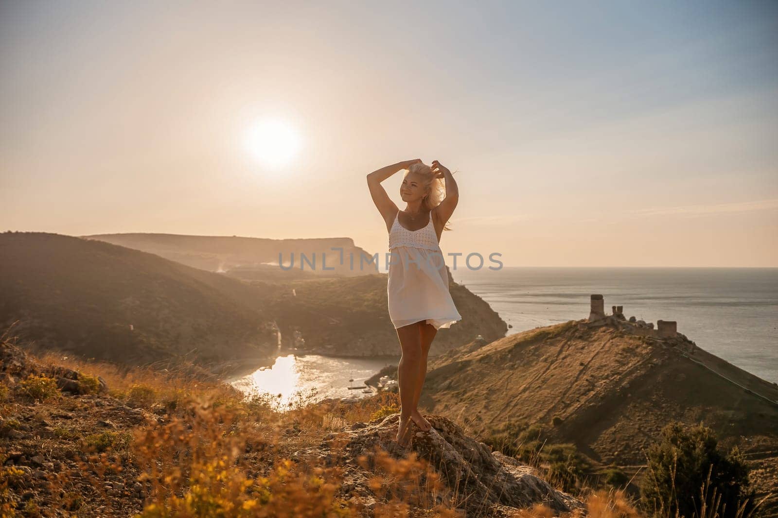 A blonde woman stands on a hill overlooking the ocean. She is wearing a white dress and she is enjoying the view. by Matiunina
