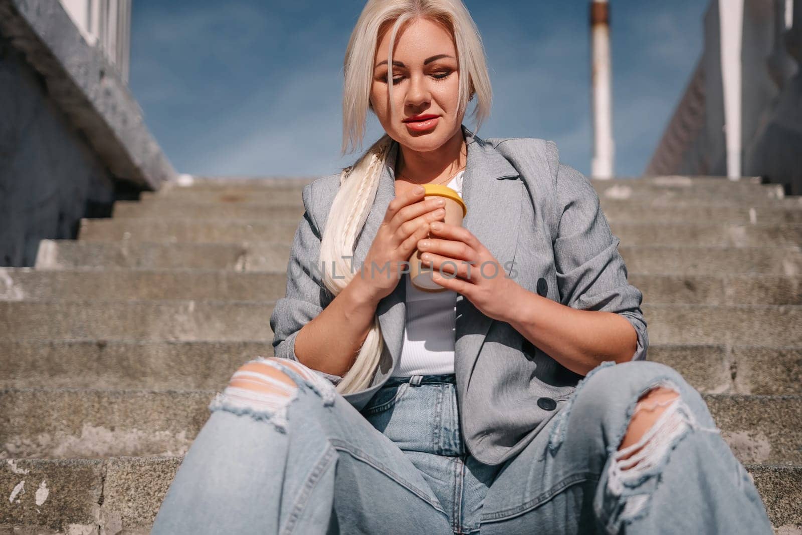 A woman in a gray jacket and jeans sits on a set of stairs with a cup in her hand
